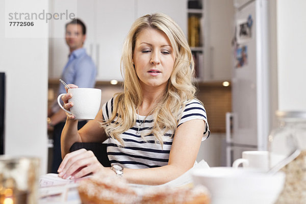 Junge Frau hält Kaffeetasse und liest Zeitung mit Mann im Hintergrund