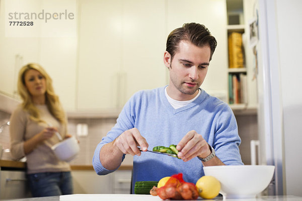 Mittlerer erwachsener Mann  der geschnittene Gurken in eine Schüssel legt  während die Frau in der Küche steht.
