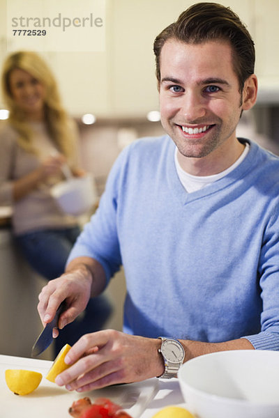 Porträt eines erwachsenen Mannes  der Zitronen an der Küchentheke mit Frau im Hintergrund schneidet.