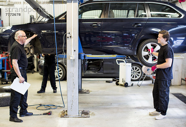 Männliche Mechaniker diskutieren im Stehen gegen ein Auto  das in der Werkstatt auf einen Wagenheber gehoben wird.