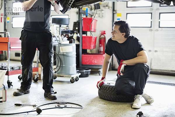 Junger Mechaniker  der in der Werkstatt auf dem Reifen sitzt und einen Mitarbeiter beim Reparieren des Autos ansieht