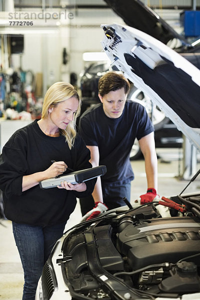 Mittlere erwachsene Mechanikerin  die ein digitales Tablett in der Hand hält  während sie mit ihrem Kollegen über den Motor des Autos diskutiert.