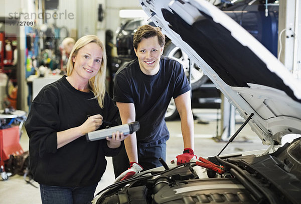 Porträt einer glücklichen erwachsenen Mechanikerin mit digitalem Tablett stehend mit Mitarbeiterin bei geöffneter Motorhaube