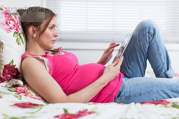 Schwangere Frau auf dem Bett liegend mit Blick auf digitales Tablett