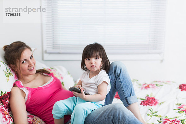 Porträt einer schwangeren Frau und einer Kleinkind-Tochter  die mit einem Smartphone auf dem Bett liegt.