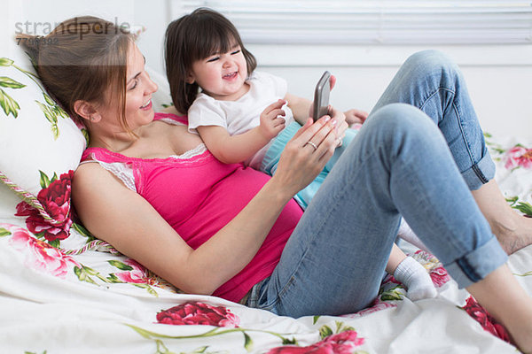 Porträt einer schwangeren Frau und einer Kleinkind-Tochter  die auf dem Bett liegt und mit dem Smartphone spielt.