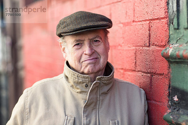 Porträt eines älteren Mannes  der sich an eine rote Ziegelwand lehnt.