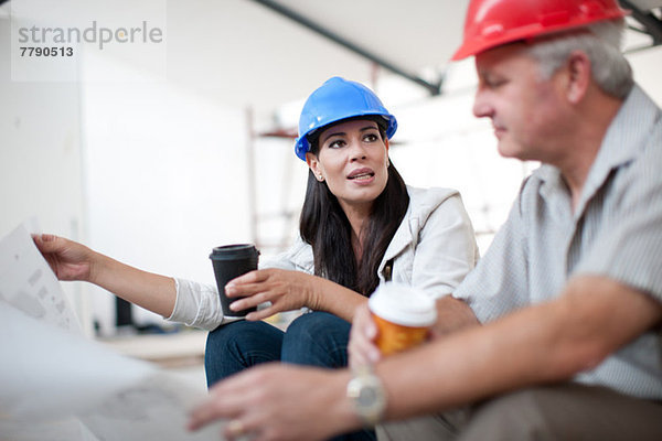 Bauarbeiterinnen und Bauarbeiter sitzen und reden und halten Kaffee und eine Blaupause.