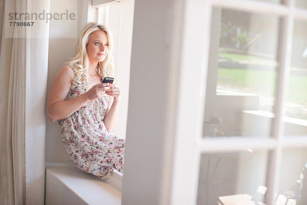 Junge Frau sitzt auf der Fensterbank und hält das Smartphone.
