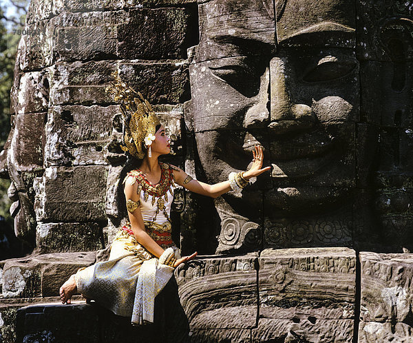 Tempeltänzerin oder Apsara sitzt vor einem Gesichter-Turm am Tempel Bayon  UNESCO-Weltkulturerbe