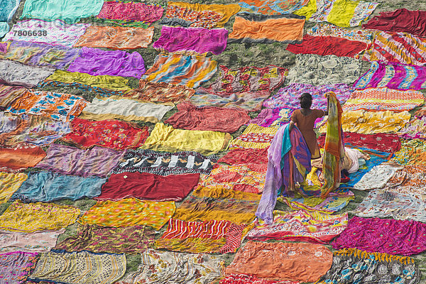 Bunte Saris sind nach dem Waschen zum Trocknen auf dem Sand ausgelegt  eine Inderin sammelt trockene Saris ein