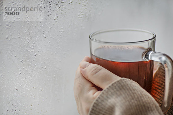 Menschliche Hände halten einen Becher Tee neben einem Fenster mit Kondenswasser.