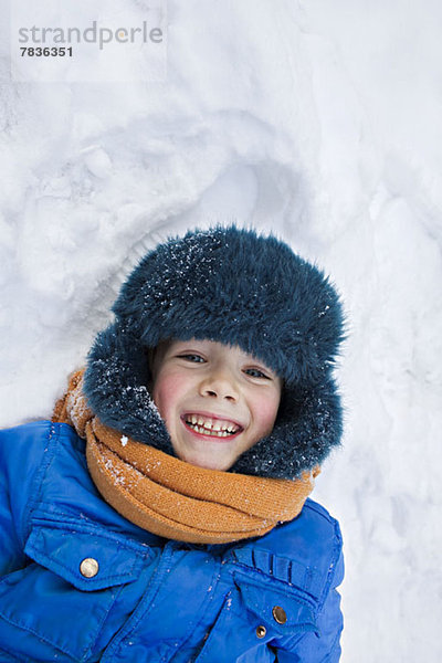 Ein junger fröhlicher Junge in warmer Kleidung draußen im Schnee liegend.
