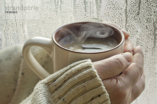 Menschliche Hände halten eine Tasse mit aufsteigendem Dampf  neben einem Fenster mit Kondenswasser.