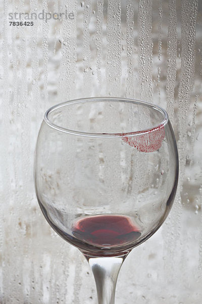 Lippenstift auf einem Glas Rotwein mit regenbedecktem Fenster im Hintergrund