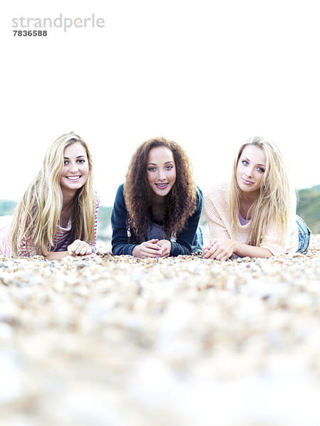 Drei fröhlich lächelnde Freunde liegen an einem felsigen Strand.