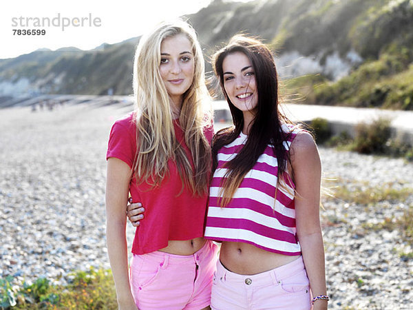 Zwei lächelnde junge Frauen  die mit den Armen umeinander am Strand stehen.
