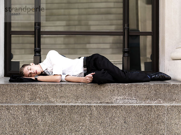 Ein junger schlafender Geschäftsmann  der auf einer Treppe vor einem Gebäude liegt.