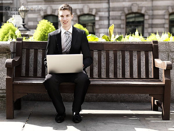 Ein fröhlicher junger Geschäftsmann  der einen Laptop benutzt  während er draußen auf einer Bank sitzt.