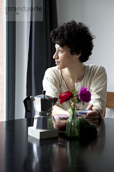 Eine Frau sitzt an einem Tisch und schaut nachdenklich aus dem Fenster.