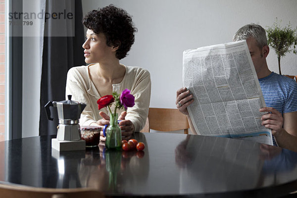 Eine ernste Frau schaut aus dem Fenster  während sich ihr Freund hinter einer Zeitung versteckt.