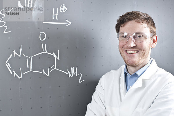 Deutschland  Porträt eines jungen Wissenschaftlers neben chemischer Gleichung auf Kreidetafel  lächelnd