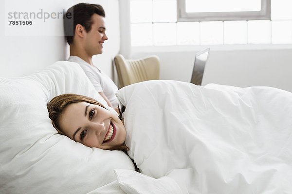 Junge Frau auf dem Bett liegend  junger Mann mit Laptop im Hintergrund