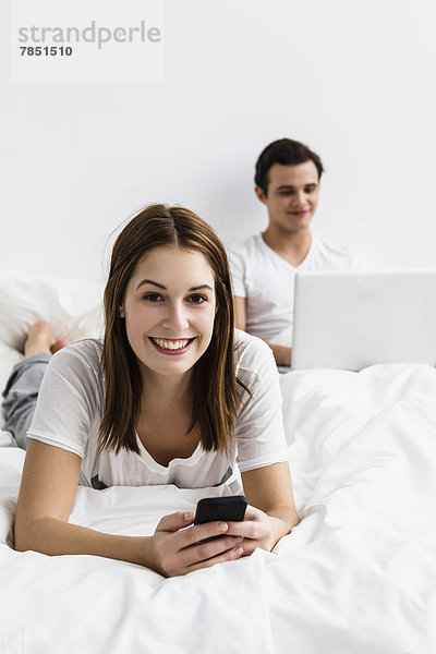 Porträt einer jungen Frau mit Handy  während ein junger Mann mit Laptop im Hintergrund lächelt.