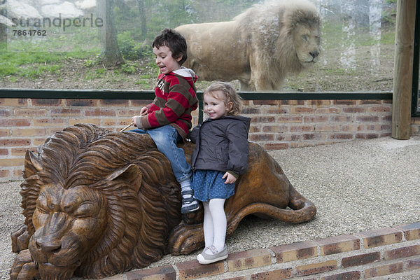 Kinder spielen auf einer Löwenstatue vor dem Löwengehege des Zoos.