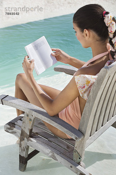 Frau sitzt auf einem Stuhl und liest ein Buch am Strand.