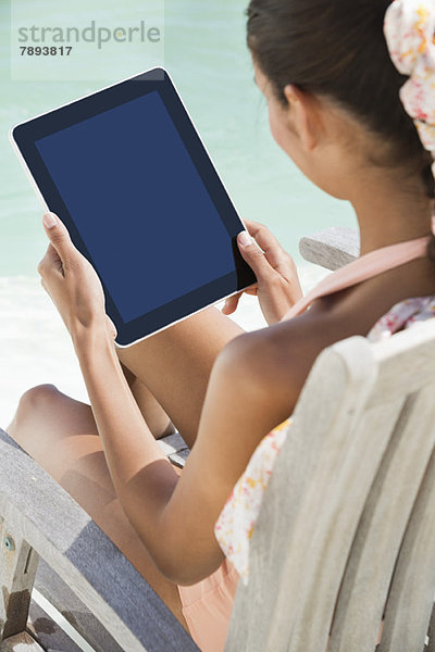 Frau sitzt auf einem Stuhl und benutzt ein digitales Tablett am Strand.