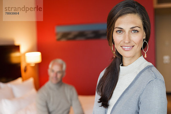 Porträt einer lächelnden Frau im Hotelzimmer mit ihrem Mann im Hintergrund