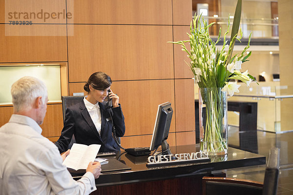 Geschäftsmann beim Lesen einer Broschüre mit einer Empfangsdame  die am Telefon an der Hotelrezeption spricht.