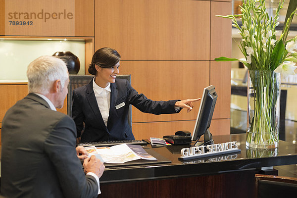 Rezeptionist  der mit einem Geschäftsmann auf einen Desktop-PC an der Hotelrezeption zeigt.