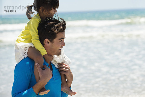Mann  der seine Tochter auf den Schultern am Strand trägt.