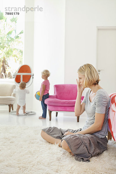 Frau sitzt auf einem Teppich und ihre Kinder spielen im Hintergrund