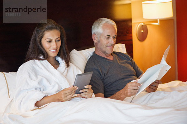 Frau  die ein digitales Tablett benutzt  während ihr Mann in einem Hotelzimmer ein Buch liest.