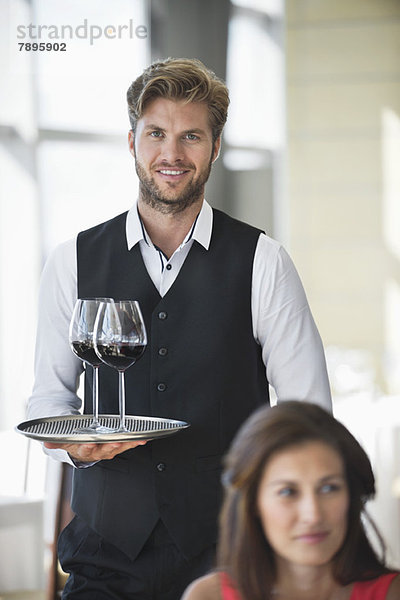Frau sitzt in einem Restaurant mit einem Kellner  der im Hintergrund ein Tablett mit Weingläsern hält.