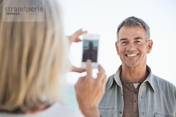 Frau beim Fotografieren ihres Mannes mit einem Handy am Strand