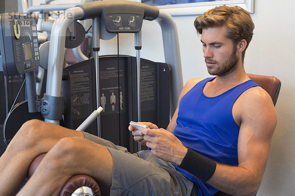 Mann  der auf einem Trainingsgerät sitzt und ein Mobiltelefon in einem Fitnessstudio benutzt.