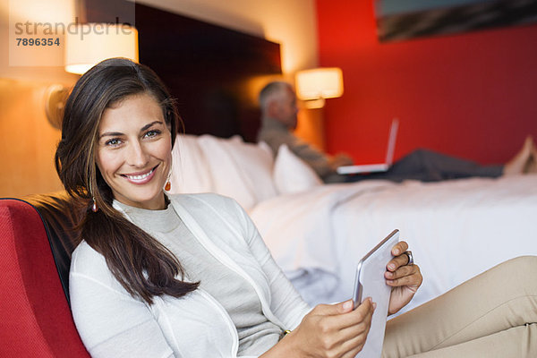 Frau mit einem digitalen Tablett im Hotelzimmer und ihr Mann mit Laptop im Hintergrund
