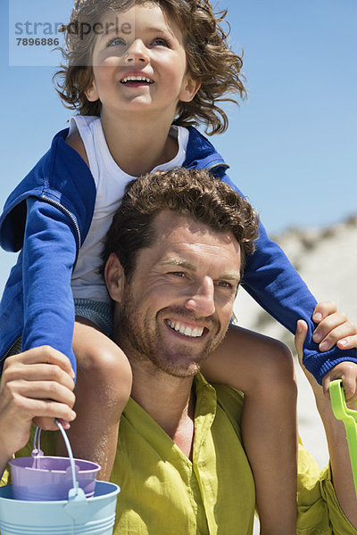 Mann trägt seinen Sohn auf den Schultern am Strand.