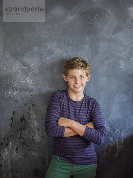 Junge lächelt mit verschränkten Armen vor einer Tafel im Klassenzimmer.