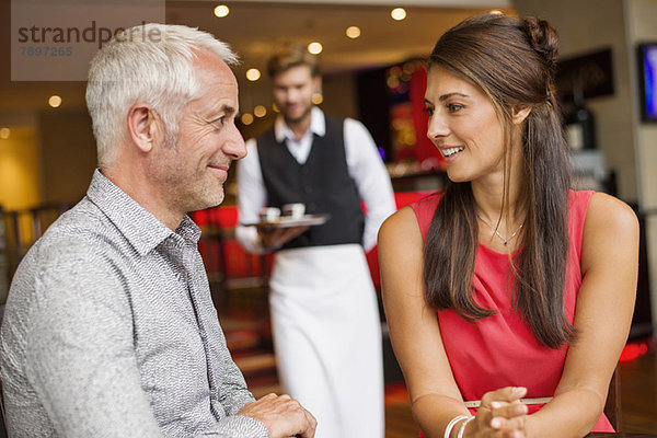 Paar lächelnd in einem Restaurant mit Kellner im Hintergrund