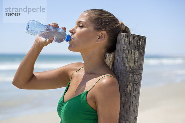 Frau lehnt sich an einen Holzpfosten am Strand und trinkt Wasser.