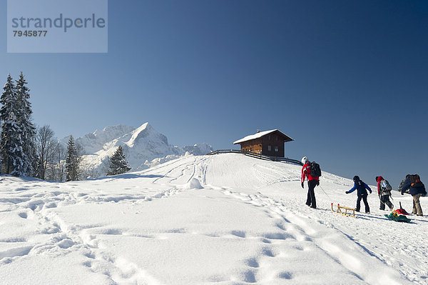 Schlittenfahrer und Holzhaus auf verschneitem Hügel  hinten die Alpspitze und Zugspitze Holzhaus