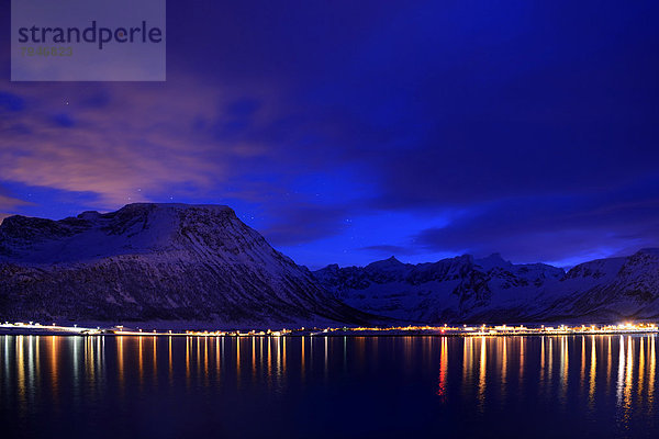 Spiegelung von Lichtern in Fjord mit Bergkette zur blauen Stunde