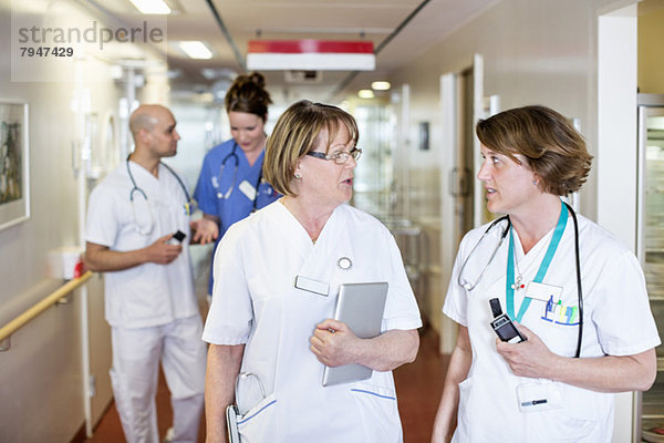 Ärztinnen diskutieren  während Kollegen im Hintergrund im Krankenhausflur sitzen