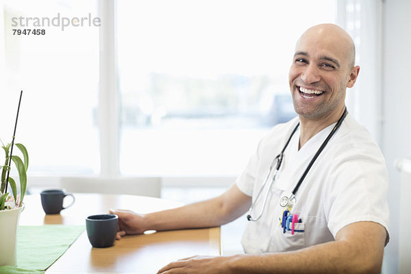 Porträt eines glücklichen Arztes mit Kaffeetasse am Schreibtisch im Krankenhaus