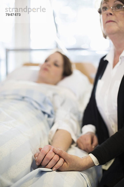 Ältere Frau schaut weg  während sie die Hand der Tochter auf dem Bett in der Krankenstation hält.